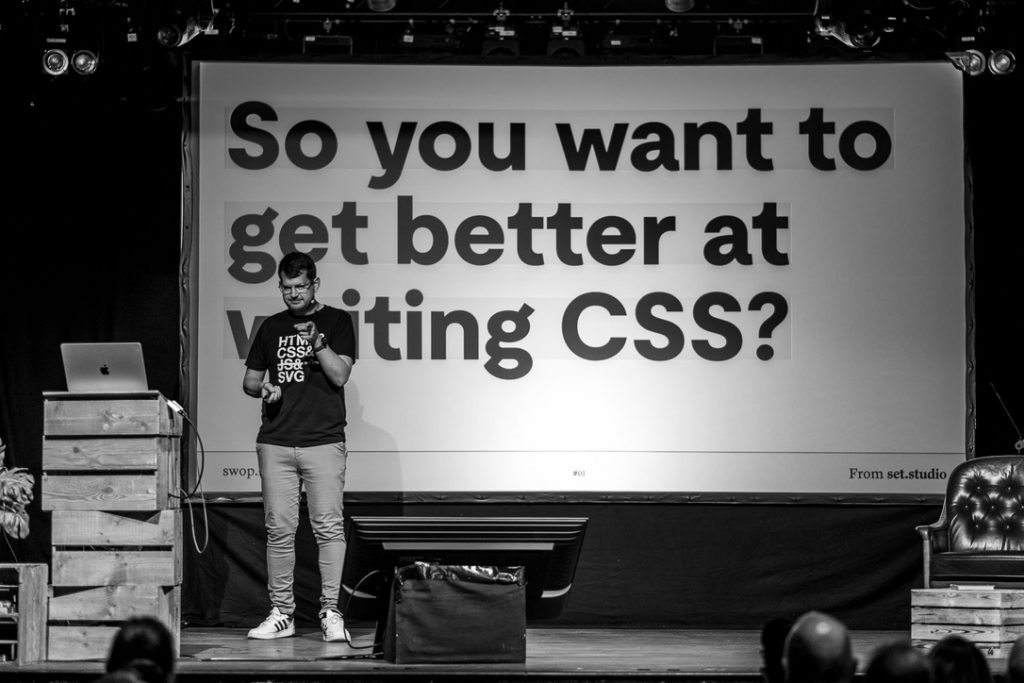 A black and white shot of me on stage. I'm pointing at the audience while the slide behind me reads: "So you want to get better at writing CSS?"